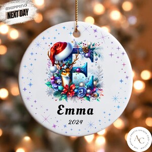 May include: A round white ceramic ornament featuring a large blue letter E decorated with a reindeer wearing a Santa hat, colourful flowers, and pine branches. The name Emma and the year 2024 are printed below the letter, surrounded by blue stars.