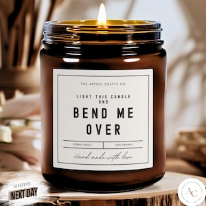 May include: A brown glass candle jar with a white label that reads "The Artful Crafts Co. Light this candle and Bend Me Over. Vegan Candle. 100% Organic. Hand made with love."