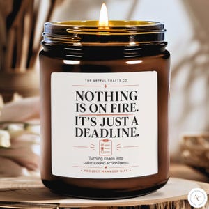 May include: A brown glass jar candle with a lit wick. The white label features the text: THE ARTFUL CRAFTS CO, NOTHING IS ON FIRE. IT'S JUST A DEADLINE. Turning chaos into color-coded action items. PROJECT MANAGER GIFT, with a checklist icon.