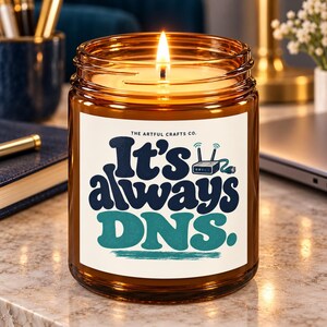 Peut inclure: Une bougie allumée dans un pot en verre ambré avec une étiquette blanche portant le texte "THE ARTFUL CRAFTS CO." et "It's always DNS." en bleu foncé et bleu sarcelle, avec un petit graphique de routeur et un câble Ethernet vert.