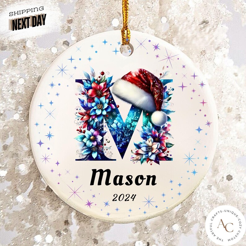 May include: A round white ceramic ornament featuring a large letter M decorated with blue, red, and purple floral patterns and a red Santa hat. The name Mason and the year 2024 are printed below the letter. The ornament has a gold hanging string.