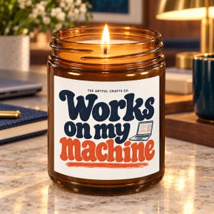 May include: A lit candle in an amber glass jar with a white label. The label features the text "THE ARTFUL CRAFTS CO." and "Works on my machine" in dark blue and orange, accompanied by a small graphic of an open laptop computer.