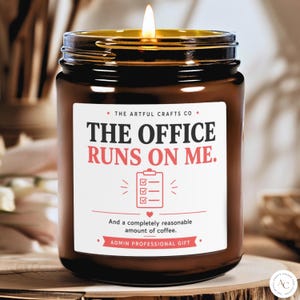 Puede incluir: Una vela en frasco de vidrio marrón con una mecha encendida. La etiqueta blanca presenta el texto THE OFFICE RUNS ON ME., un icono de portapapeles, el texto And a completely reasonable amount of coffee. y ADMIN PROFESSIONAL GIFT.
