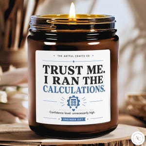 May include: A brown glass jar candle with a lit wick and a white label. The label reads: THE ARTFUL CRAFTS CO, TRUST ME. I RAN THE CALCULATIONS., a calculator icon, Confidence level: unnecessarily high., and ENGINEER GIFT.