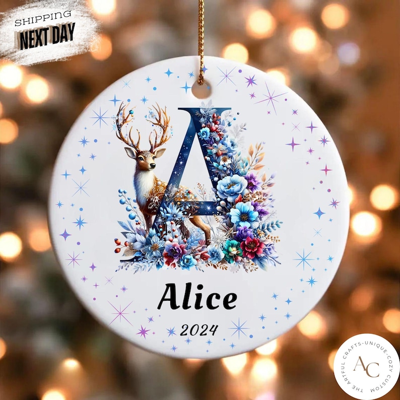 May include: A round white ceramic ornament featuring a large dark blue letter A decorated with a deer and colourful floral patterns. The name Alice and the year 2024 are printed below the letter. The ornament is accented with small blue stars.
