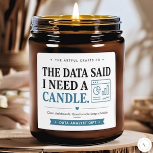 May include: A brown glass jar candle with a lit wick and a white label. The label reads: THE ARTFUL CRAFTS CO, THE DATA SAID I NEED A CANDLE, with a chart icon, a heart, and the text: Clean dashboards. Questionable sleep schedule. DATA ANALYST GIFT.