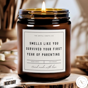 May include: A brown glass candle jar with a white label that reads "Smells like you survived your first year of parenting". The label also says "Vegan Candle" and "100% Organic". The candle is lit and the flame is visible.