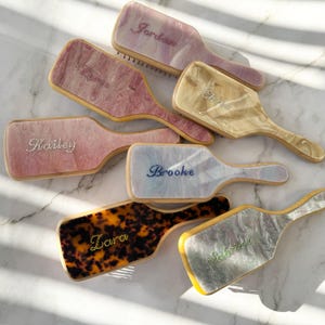 Könnte beinhalten: Eine Auswahl personalisierter Haarbürsten mit Namen wie "Jordan" und "Brooke". Die Bürstenköpfe weisen Marmor- und Schildpattmuster auf. Die Griffe sind aus Holz. Die Schriftzüge auf den Bürsten sind dekorativ.