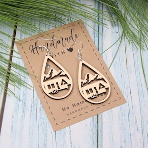 May include: Wooden teardrop earrings with a mountain and camper design. The earrings are light brown with silver-colored ear wires. The earrings are displayed on a brown card with the words "Handmade with" and "No Name J.B." printed on it.