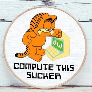 May include: A circular wooden embroidery hoop containing a cross-stitch design of an orange, pixelated cat standing next to a yellow computer. The screen displays the letters OW in white. Below the cat, the text reads COMPUTE THIS SUCKER.