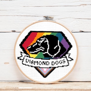 May include: A cross-stitch embroidery hoop featuring a rainbow-coloured diamond shape containing a black silhouette of a dog's head. The diamond shape has a banner underneath that reads "DIAMOND DOGS".
