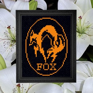 May include: Framed cross-stitch art featuring an orange fox design within an oval on a black background. The word "FOX" is stitched below the fox. The frame is black. The background is white flowers.