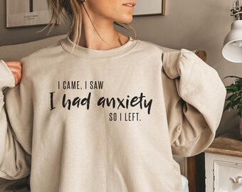 May include: A beige sweatshirt with the text "I came, I saw, I had anxiety, so I left." printed in black.