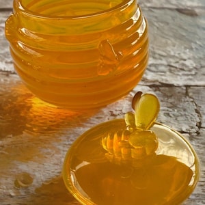 Puede incluir: Un tarro de miel de vidrio transparente con tapa a juego. El tarro tiene forma cilíndrica con estrías horizontales y asas en forma de abeja. La tapa tiene un asa en forma de abeja. El vidrio es de color ámbar cálido.