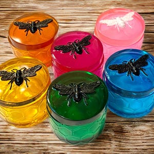 Op de afbeelding: Een verzameling van zes kleurrijke, decoratieve potten met bijvormige deksels. De potten zijn in tinten oranje, roze, geel, groen, blauw en magenta. De deksels hebben een gedetailleerd bijenontwerp.