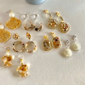 Op de afbeelding: Assortiment van gouden en zilveren oorbellen met verschillende schelp- en harsontwerpen. De oorbellen hebben verschillende vormen, waaronder cirkels, ovalen en rechthoeken. Sommige oorbellen hebben een gouden afwerking, terwijl andere een zilveren afwerking hebben.