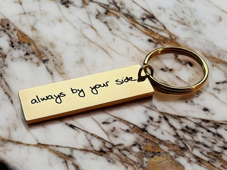 Op de afbeelding: Gouden sleutelhanger met een rechthoekig label waarop in handschrift "always by your side" staat.