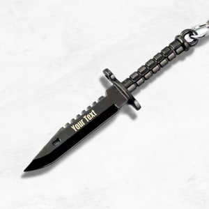 Puede incluir: Un llavero de metal negro con forma de cuchillo con una hoja dentada. La hoja tiene el texto "Your Text" grabado.