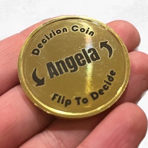 May include: Gold-colored coin with the text "Decision Coin" and "Flip To Decide" around the edge. The center of the coin has an arrow pointing to the right and the word "Angela" written in a curved line.