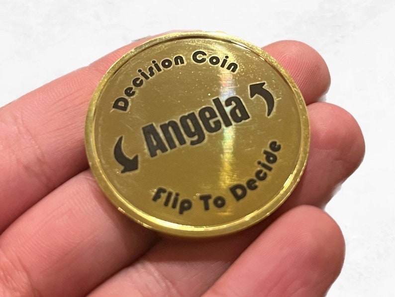 May include: Gold-colored coin with the text "Decision Coin" and "Flip To Decide" around the edge. The center of the coin has an arrow pointing to the right and the word "Angela" written in a curved line.