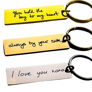 Op de afbeelding: Drie gouden en zilveren sleutelhangers met handgeschreven berichten. De eerste sleutelhanger zegt "You hold the key to my heart". De tweede sleutelhanger zegt "always by your side". De derde sleutelhanger zegt "I love you xoxo".