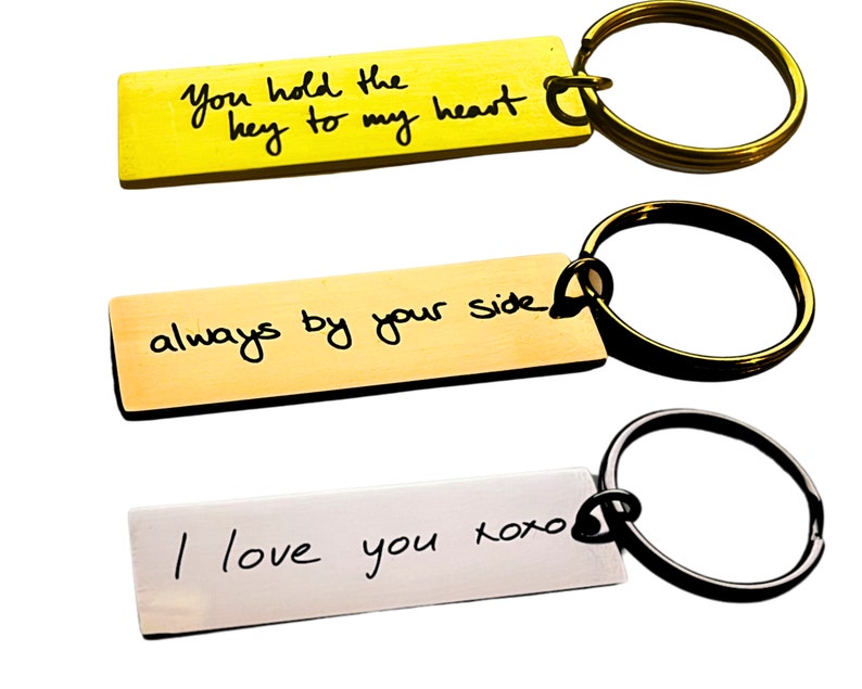 Op de afbeelding: Drie gouden en zilveren sleutelhangers met handgeschreven berichten. De eerste sleutelhanger zegt "You hold the key to my heart". De tweede sleutelhanger zegt "always by your side". De derde sleutelhanger zegt "I love you xoxo".