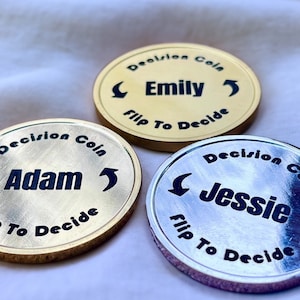 May include: Three gold-colored decision coins with black lettering. The coins feature the names "Adam", "Emily", and "Jessie" with arrows pointing in opposite directions. The text "Decision Coin" and "Flip To Decide" are also printed on each coin.