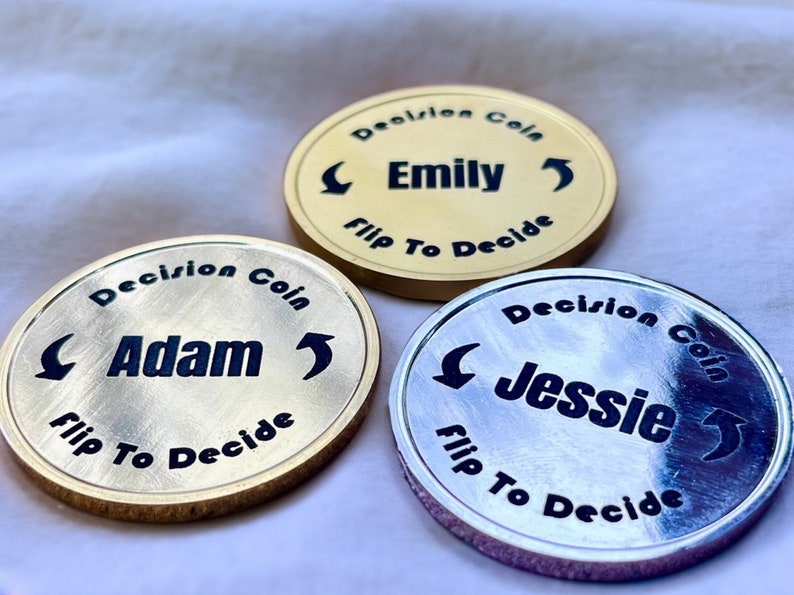 May include: Three gold-colored decision coins with black lettering. The coins feature the names "Adam", "Emily", and "Jessie" with arrows pointing in opposite directions. The text "Decision Coin" and "Flip To Decide" are also printed on each coin.