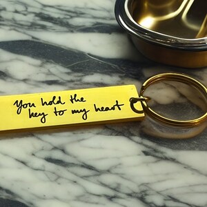 Op de afbeelding: Een gouden sleutelhanger met een rechthoekig label waarop staat "You hold the key to my heart".