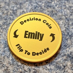 May include: Gold-colored coin with the text "Decision Coin" and "Flip To Decide" around the edge. The center of the coin has the name "Emily" and arrows pointing left and right.