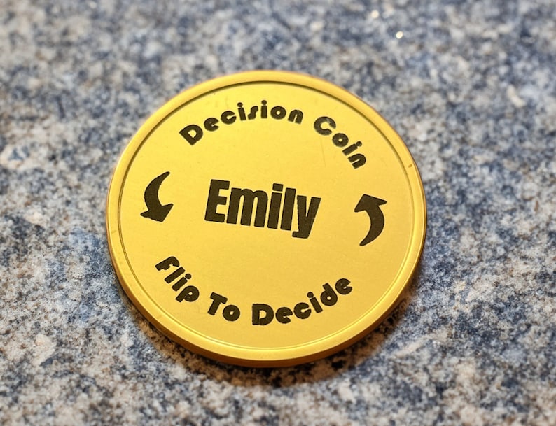 May include: Gold-colored coin with the text "Decision Coin" and "Flip To Decide" around the edge. The center of the coin has the name "Emily" and arrows pointing left and right.