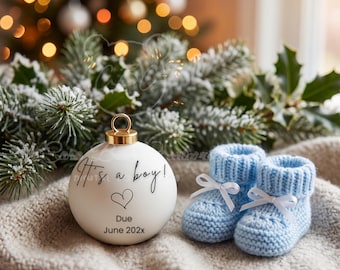May include: A white Christmas ornament with gold accents and the text "It's a boy!" and "Due June 202x". Next to the ornament are two light blue knitted baby booties with white bows. The background includes a Christmas tree.