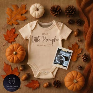 Puede incluir: Un body de bebé color crema con el texto "Our Little Pumpkin October 202X" está rodeado de decoraciones otoñales. Incluyen calabazas naranjas, hojas de otoño, piñas, bellotas y una imagen de ultrasonido.
