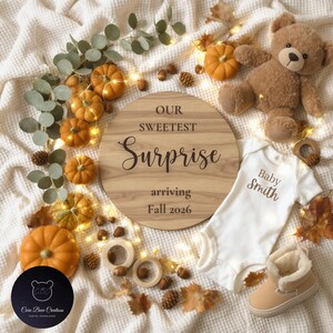 May include: A wooden sign with the text "OUR SWEETEST Surprise arriving Fall 2026" is surrounded by pumpkins, leaves, and a teddy bear. A white baby onesie and tan baby boots are also present, along with wooden rings and pinecones.