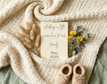 May include: A cream-colored, chunky knit blanket with a card that reads "Adding a little more love to our family May 2026." The card is surrounded by dried flowers and greenery. Two pairs of beige baby booties with bows are at the bottom.