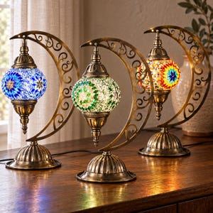 Puede incluir: Tres lámparas de mesa con globos de cristal de mosaico en patrones azules, verdes y multicolores. Cada lámpara cuelga de un soporte de metal color latón con forma de media luna, con recortes de volutas y una base redonda.