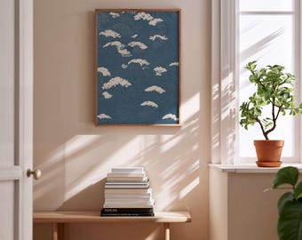 May include: A framed art print featuring a blue and white cloud design. The artwork is displayed on a wall above a wooden bench with a stack of books. A potted plant sits on a windowsill, and sunlight streams through the window.