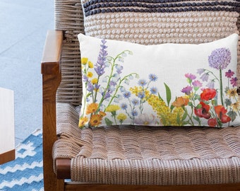 May include: A rectangular decorative pillow with a floral design featuring colorful wildflowers, including red poppies, purple alliums, and yellow blooms. The pillow is on a woven chair with a cream-colored pillow with a dark stripe.