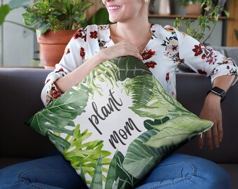 May include: A white cushion with a green tropical leaf pattern and the text "plant mom" in black lettering.