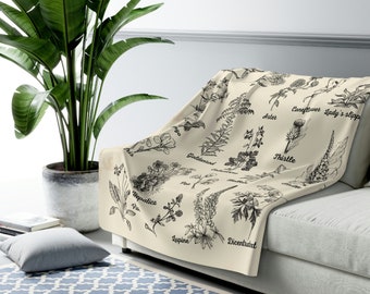 May include: A cream-coloured throw blanket draped over a sofa, featuring detailed black botanical illustrations of various flowers and plants. The names of the plants are printed in a classic font. The blanket is a decorative home textile.