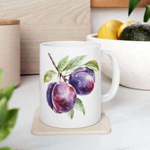 May include: White ceramic mug with a watercolor illustration of purple plums on a branch with green leaves.