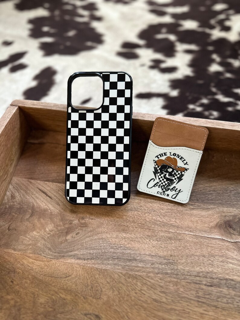 May include: Black and white checkered phone case with a black border. The case is next to a brown leather card holder with a white skull graphic and the text "The Lonely Cowboy Club".