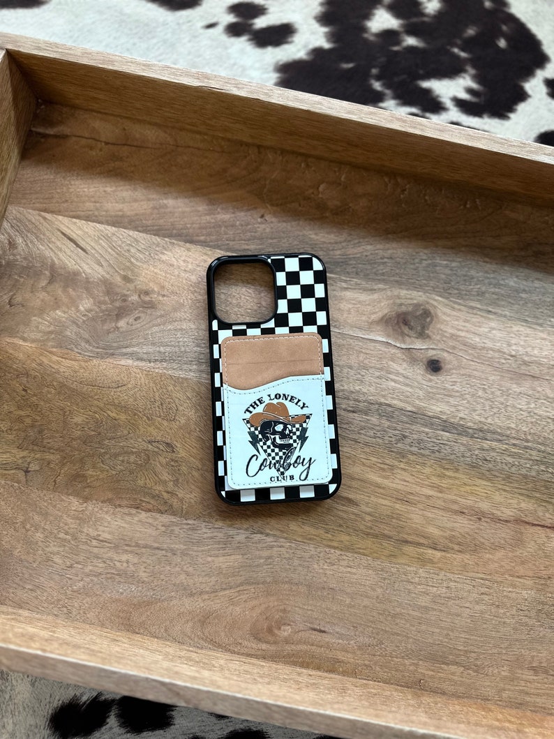 May include: A black and white checkered phone case with a brown leather pocket and a skull graphic that says "The Lonely Cowboy Club".