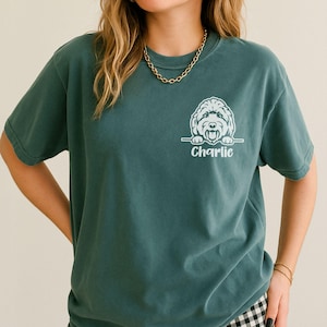 Op de afbeelding: Een blauwspar-kleurig t-shirt met korte mouwen en een witte afbeelding van een hondengezicht op de linkerborst. Onder de hondenillustratie staat de naam Charlie gedrukt in een wit cursief lettertype. Het shirt heeft een ronde hals.