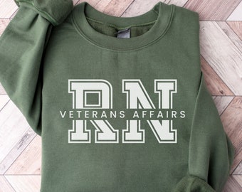 May include: An olive green sweatshirt featuring the white text "RN VETERANS AFFAIRS". The sweatshirt has a crew neck and long sleeves with ribbed cuffs. The garment is made of a soft material, suitable for casual wear.