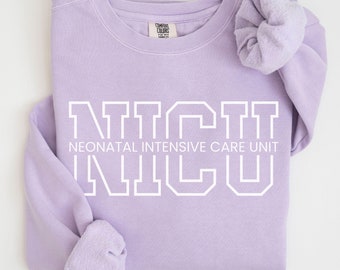 May include: A lavender sweatshirt with the text "NICU" in white, with the words "Neonatal Intensive Care Unit" underneath. The sweatshirt is folded in half and a lavender scrunchie is on top of the folded sleeve.