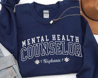 May include: A navy blue crewneck sweatshirt featuring the white text MENTAL HEALTH COUNSELOR in a collegiate font, with the name Stephanie in script below, flanked by two small white daisy icons. A gold chain necklace rests on the collar.