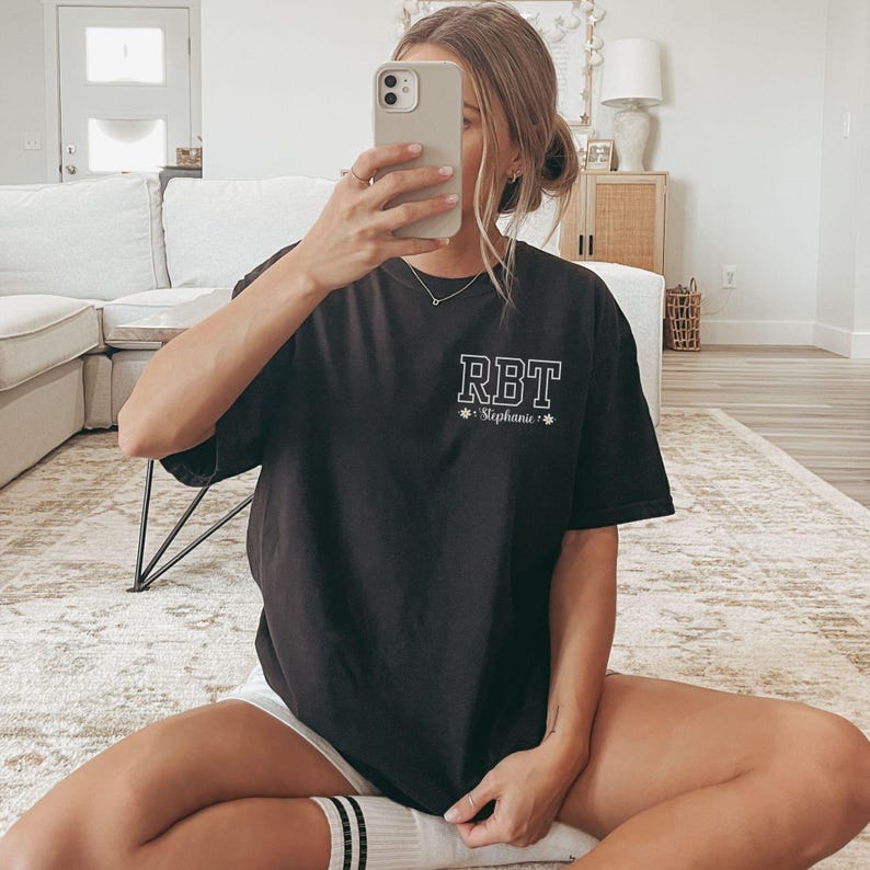 May include: Black short-sleeved t-shirt with the letters "RBT" in a white, block font on the upper left chest. Below the letters is the name "Stephanie" in a smaller font. The shirt is worn with white shorts.