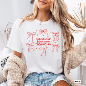 May include: A white short-sleeved t-shirt featuring red line art of six decorative bows arranged around the centred red text that reads REGISTERED BEHAVIOR TECHNICIAN. The design includes small four-pointed star accents between the bows.