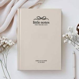 May include: A beige hardcover notebook with the text "little notes FOR THE BIG DAY" and a bow graphic. The names "AMELIA & JOHN" and the date "12.29.2025" are printed at the bottom. The notebook is surrounded by white flowers and a pen.